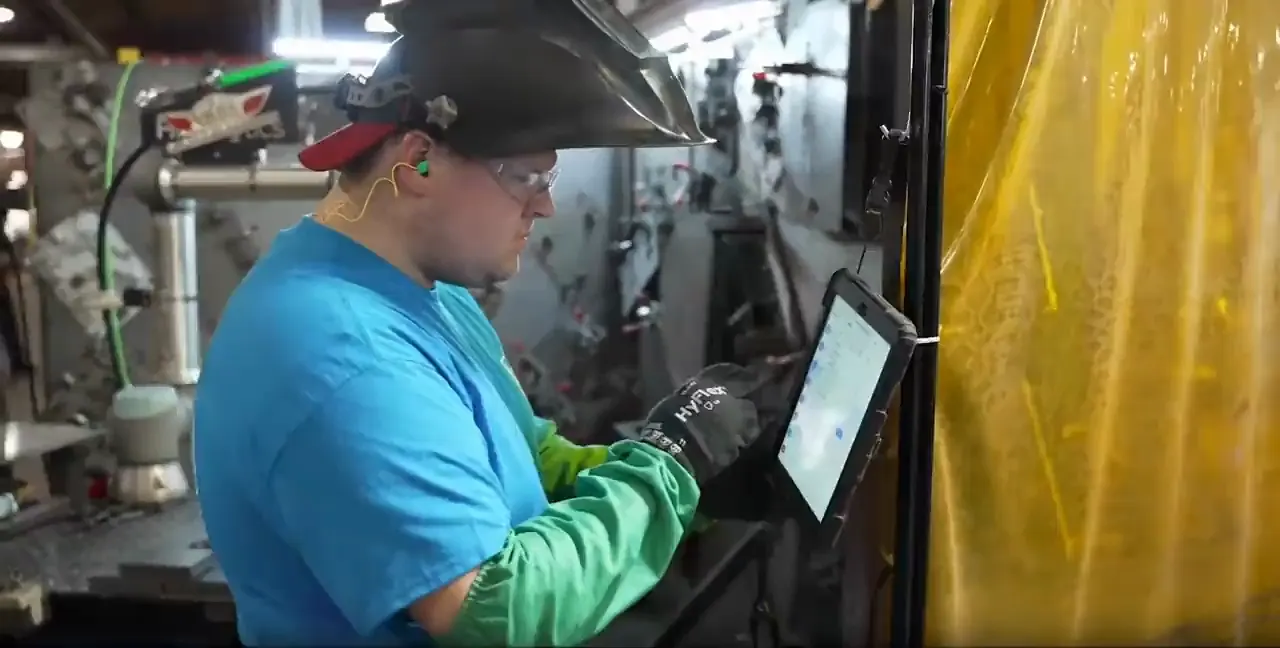 Operator in a welding helmet setting up a part on a tablet beside a cobot cell